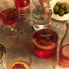 A glass bottle pours a dark red cocktail spirit into a glass of red drink on a marble countertop, with other glasses, a bottle, and green olives in the background.
